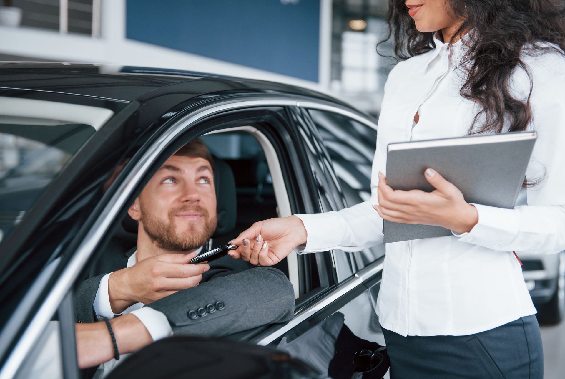 Herzlichen Glückwunsch zum Neukauf. Glücklicher Besitzer von neuen Auto sitzt im Inneren und nimmt Schlüssel von weiblichen Manager